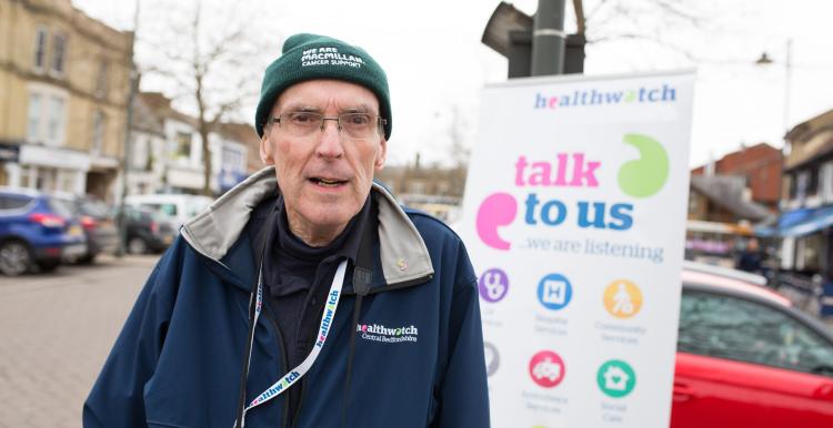 Healthwatch volunteer standing infront of a Healthwatch sign saying 'talk to us'. 
