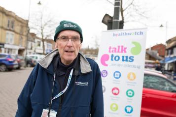 Healthwatch volunteer standing infront of a Healthwatch sign saying 'talk to us'. 